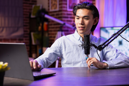 Asian male content creator records podcast episode in professional home studio. Young male adult at desk with microphone and laptop, creating digital content in cozy, modern recording setup.