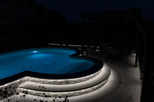 A modern outdoor pool glowing at night with vibrant LED lighting inside and around the water, creating a luxurious, serene, and atmospheric scene ideal for relaxation and nightlife visuals.