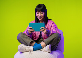 Smiling Asian Gen Z woman using a digital tablet while sitting cross-legged against a vibrant neon green background.