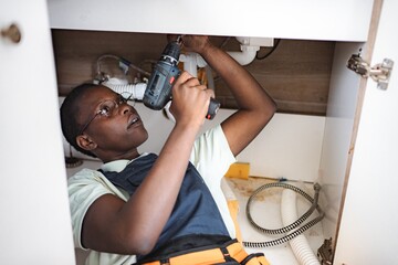 Young female plumber lying down and using electric screwdriver beneath kitchen sink