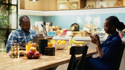Happy couple looking at something on tablet enjoying breakfast together, sitting at the kitchen table. Drinking coffee and chatting over fresh croissants for a relaxing morning routine. Camera B. - Powered by Adobe