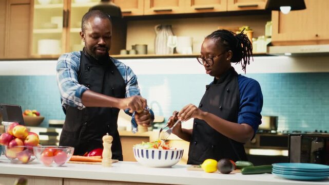 Black boyfriend seasoning salad with pepper and girlfriend stopping him, enough spices or condiments for healthy lunch in bowl. Joyous couple with aprons preparing meal together. Camera B.
