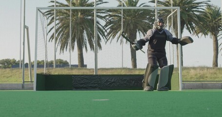 Fototapeta premium Standing teen field hockey goalkeeper wearing helmet and pads, holding stick, guarding goal on turf