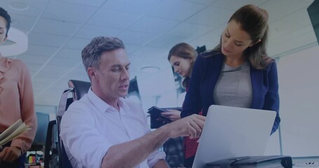 Pointing seated mature man gesturing toward laptop at office desk, colleagues leaning in blazers