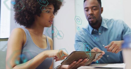 Reviewing colleagues examining tablet with pen in meeting room, sleeveless, blue shirt, hexagon UI