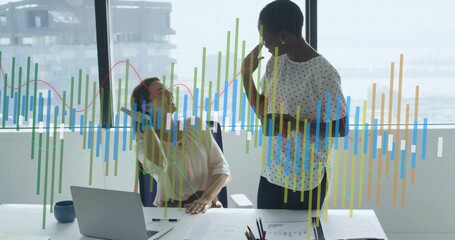 Reviewing two office women in blouses analyzing charts at bright office desk with laptop and data