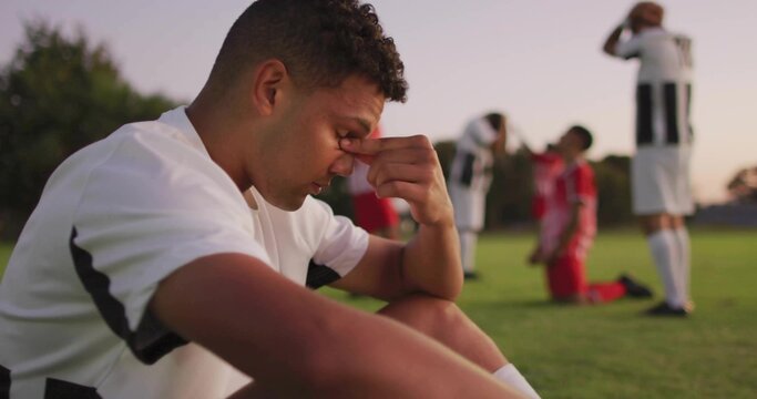Sitting soccer player pinching nose on pitch at dusk, wearing white kit, knee-high socks and cleats