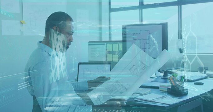 Examining architect in light-blue shirt and glasses, reviewing blueprints at office with monitors - Powered by Adobe