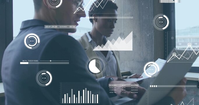 Reviewing laptop charts, two colleagues in suit and light blazer with documents by office window
