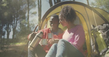 Pouring couple in red jacket and pink top sitting at yellow tent, thermos and camping mugs