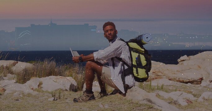 Sitting male hiker checking tablet on coastal rocks at dusk, shorts, sandals, backpack, rolled-mat