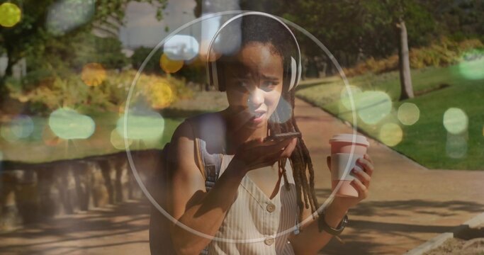 Talking female with headphones, backpack, speaking into phone and holding coffee on park path