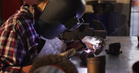 Welding welder using torch, feeding filler rod at metal shop bench, wearing helmet, apron, gloves