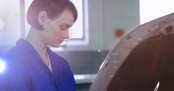 Inspecting woman technician in blue coveralls leaning over car with open hood in garage, lens flare - Powered by Adobe