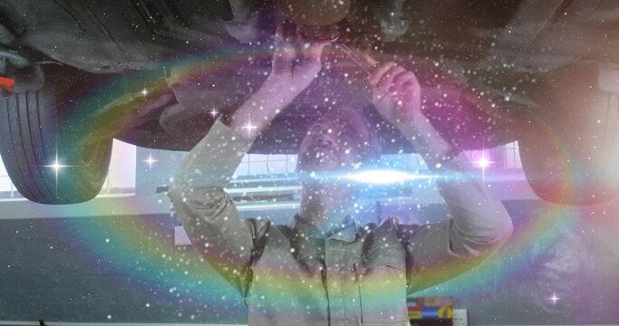 Fixing mechanic in collared shirt reaching under vehicle on lift in bay with tire, rainbow ring - Powered by Adobe