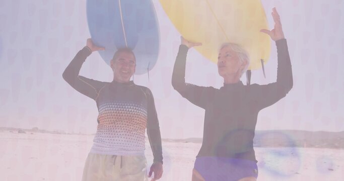 Standing couple wearing rash guards and swim bottoms carrying blue and yellow surfboards on beach