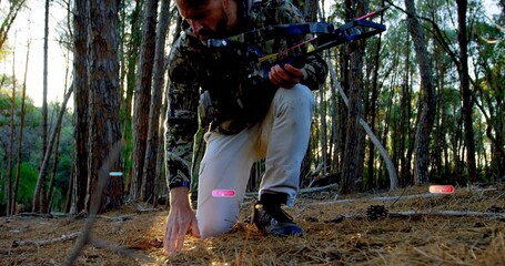Fototapeta premium Kneeling adult hunter scanning pine forest floor, holding compound bow and wearing camo coat