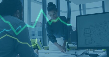 Pointing woman architect wearing dark top and light skirt leaning at office table, with plans, vest