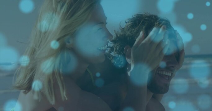 Giving woman covering man's eyes carrying him at beach shoreline, swimsuit top, trunks, blue bokeh