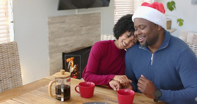 Diverse couple leaning at home by fireplace, reaching for red mugs and sipping coffee for warmth