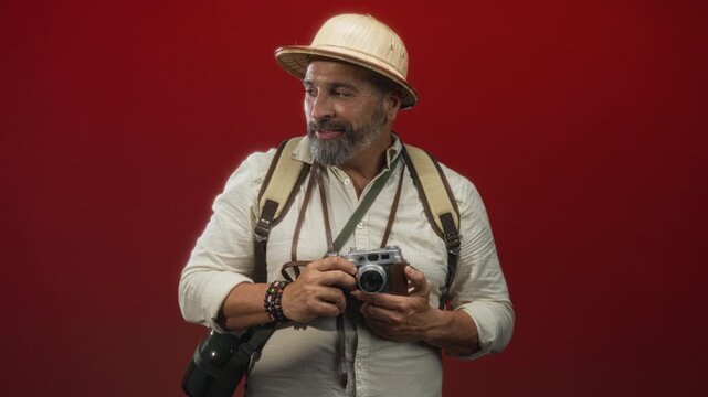Man holding a vintage camera and wearing a pith helmet and backpack in studio with red backdrop; adventure curiosity.