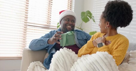Diverse couple with man wearing Santa hat surprising partner with green gift on couch at home - Powered by Adobe