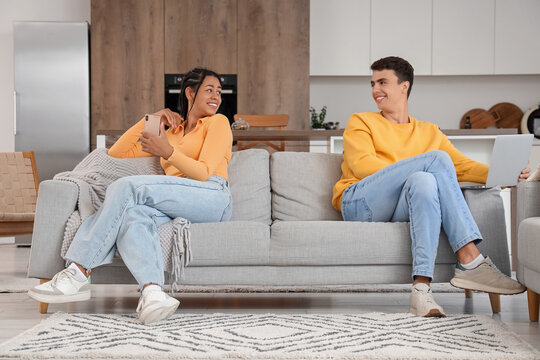 Young couple using mobile phone and laptop on sofa at home