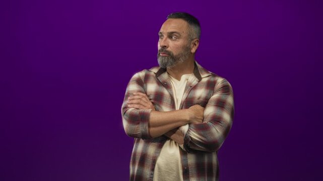 Middle aged man with crossed arms and visible beard wearing a plaid shirt in purple studio; contemplation.