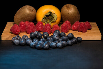 Kiwi con caqui arandanos y frambuesas sobre tabla de madera y fondo negro
