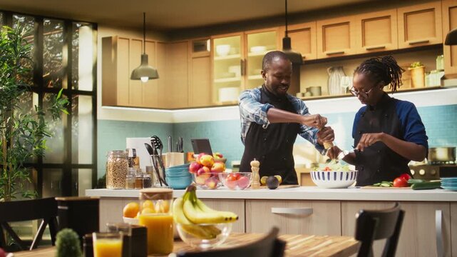African american man seasoning the salad with mix of spices and pepper, pouring condiments for a tasty vegetarian meal. Smiling woman mixing fresh vegetables together, healthy lifestyle. Camera A.
