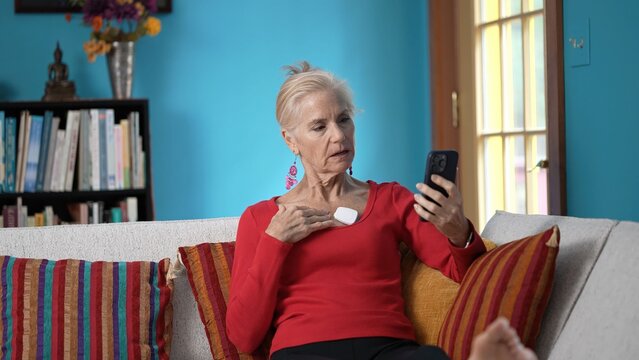 Mature woman wearing a heart monitor engages with her smartphone while sitting comfortably on a couch at home