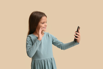 Cute little happy girl with mobile phone waving hand while video chatting on beige background