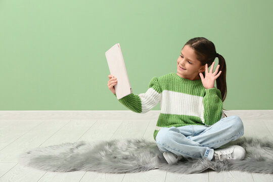 Cute little happy girl with tablet waving hand while video chatting on carpet near green wall