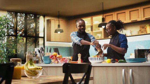 Black boyfriend seasoning salad with pepper and girlfriend stopping him, enough spices or condiments for healthy lunch in bowl. Joyous couple with aprons preparing meal together. Camera A.