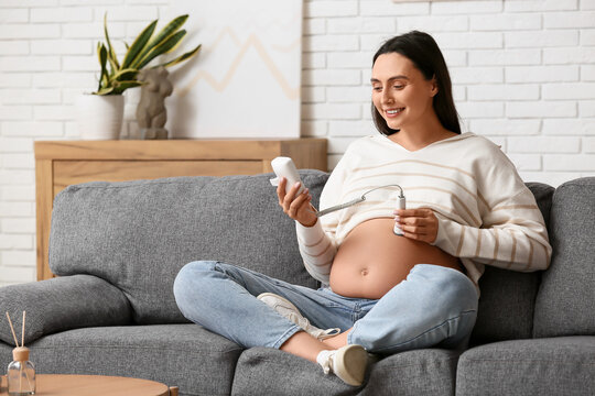 Young pregnant woman with fetal doppler listening to baby's heartbeat on sofa at home
