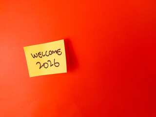 The image shows a yellow sticky note with the handwritten message WELCOME 2026 on a bright orange background