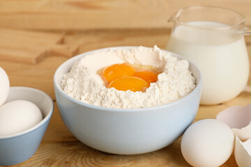 Bowl of flour with fresh chicken eggs and jug of milk on wooden table