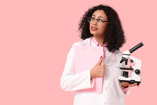 Female African-American doctor with microscope and clipboard on pink background