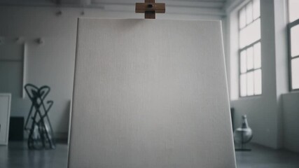 A large, blank white canvas stands on an easel in a bright, minimalist art studio with large windows. Other art supplies are visible in the background.
