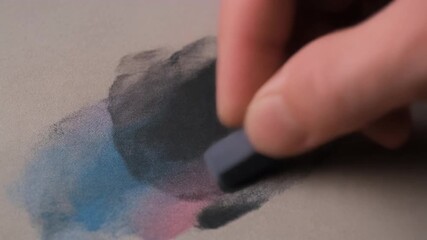 A hand applies dark pastel chalk to paper, blending with existing blue and pink colors in an artistic process.