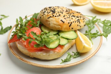 Delicious bagel with salmon, cucumber, arugula and lemon on white table, closeup