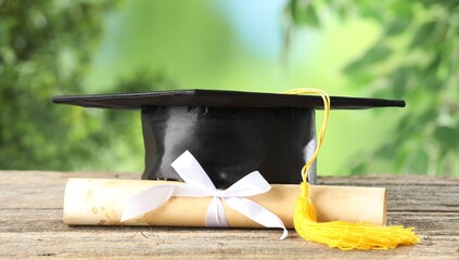 Graduation cap and diploma on wooden table against blurred background, closeup