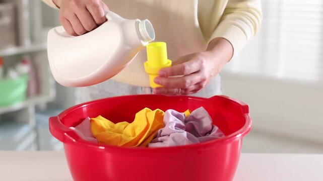 Woman pouring detergent into plastic basin with clothes at table indoors, closeup. Camera moving forward