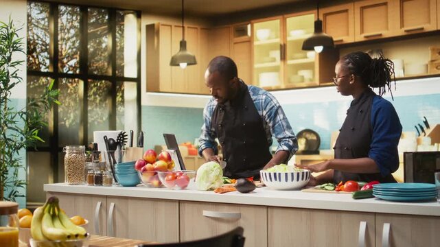 African american guy adding chopped vegetables from the cutting board, pushing slices of carrots, cucumbers and bell peppers in the salad bowl. Man and woman preparing a vegetarian lunch. Camera A.
