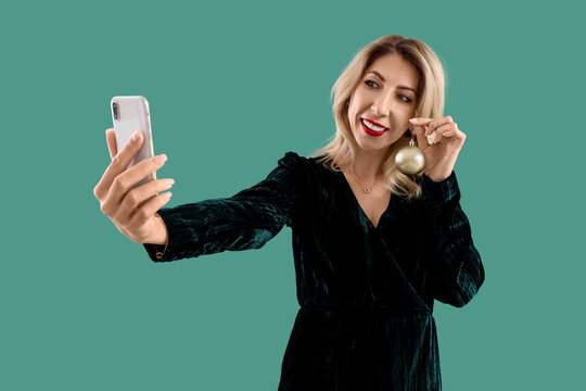 Beautiful middle-aged woman with Christmas bauble taking selfie on green background