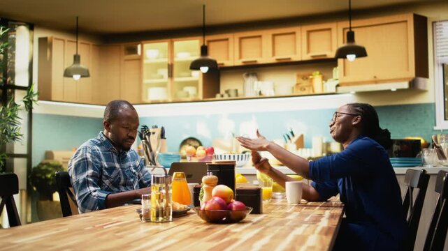 African american man working too hard and woman asking for a break, showing him the timeout sign and closing his laptop. Workaholic boyfriend ignoring his girlfriend during breakfast. Camera A.