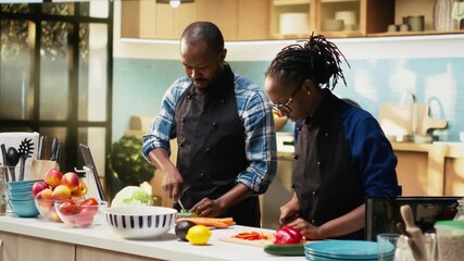 Black young lovers chopping veggies and preparing ingredients on the cutting board, cooking together. Couple cutting organic vegetables for a salad bowl, relaxed fun energy at home. Camera B. - Powered by Adobe