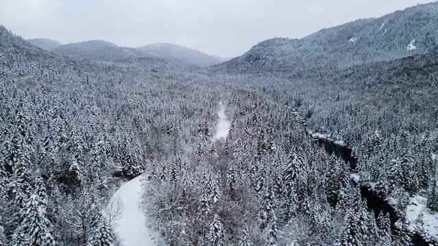 Aerial view of a snow-covered road crossing the snow-covered boreal forest and heading towards the mountains on a winter day.