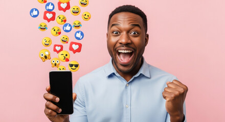 An excited African American man holds a smartphone displaying emojis over a pink backdrop, celebrating his online engagement and social media success in studio light