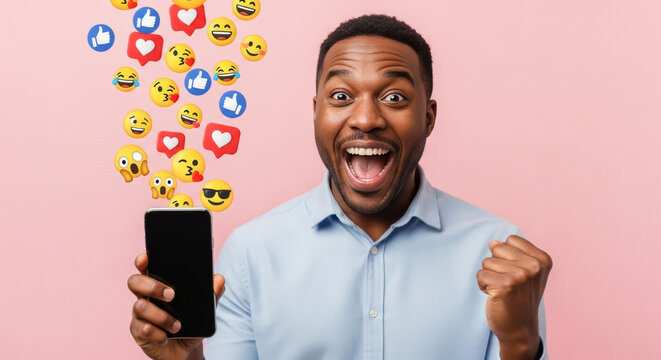 An excited African American man holds a smartphone displaying emojis over a pink backdrop, celebrating his online engagement and social media success in studio light